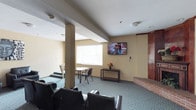 The TV Room at 2525 Highland Ave in National City is a single-floor communal lounge designed for comfortable gathering and relaxation. The room features a wall-mounted flat-screen television as its central focal point, positioned at an ideal viewing height for the seating area arranged in front of it. Black cushioned chairs provide ample seating, creating a casual and inviting environment.

The space is finished with patterned carpet flooring in a muted teal tone that adds a subtle decorative touch underfoot. Warm yellow-toned walls contribute to a welcoming ambiance throughout. A structural column is integrated into the layout, adding architectural character to the room.

Natural light filters in through a window fitted with horizontal blinds, brightening the space during the day. A small table with chairs offers an additional area for more focused activities such as reading or working. A narrow console table along the far wall holds a floral arrangement, adding a modest decorative accent. A brick-accented element along one wall provides a hint of textural contrast to the otherwise smooth interior finishes. Ceiling-mounted lighting and safety fixtures complete the practical, well-maintained environment, making this TV room a functional and comfortable communal space.