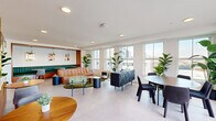 Located at 601 N Broad Street in Philadelphia's vibrant Northern Liberties neighborhood, this thoughtfully designed communal space offers a sophisticated blend of lounge and dining functionality across a single, open-plan floor. Large windows line the perimeter, flooding the interior with abundant natural light and framing sweeping views of the Philadelphia skyline. Light-toned porcelain tile flooring runs throughout, lending a clean and modern foundation to the warm, eclectic décor.

A standout architectural feature is the curved, fluted-wood bar with a white marble countertop, accented with brass hardware and a suspended overhead shelving unit adorned with lush trailing plants. This central element elegantly divides the space while maintaining an open, airy feel. The lounge area features a tufted cognac leather sofa, an organic-shaped green area rug, and an array of accent seating arranged around low tables. A built-in banquette upholstered in deep teal channeled leather anchors a cozy dining nook, illuminated by gold pendant lights and framed by colorful artwork. The dedicated dining area includes a round table with modern black chairs and direct access to an outdoor terrace through sliding glass doors, seamlessly connecting the interior to the exterior.