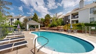 Located at 3308 Quail Lake Rd in Colorado Springs, Colorado, this outdoor amenity area offers a well-appointed communal retreat set against a backdrop of mature trees and multi-story residential buildings. The centerpiece of the space is a generously sized, kidney-shaped swimming pool with clear blue water, accessible via stainless steel entry rails and steps. The surrounding concrete pool deck is expansive, providing ample room for relaxation and movement. Numerous lounge chairs and chaise lounges are arranged throughout the deck, inviting residents to unwind and soak up the Colorado sun. A covered pavilion with a pitched shingle roof anchors one end of the space, sheltering additional seating and gathering areas ideal for socializing in the shade. Umbrella-shaded tables and chairs are distributed around the perimeter for casual outdoor dining. The area is enclosed by sleek black metal fencing, offering both safety and an unobstructed view of the surrounding greenery. Lush landscaping, including mature deciduous trees and shrubs, frames the space and provides a natural, park-like atmosphere. The combination of open sky, vibrant foliage, and well-maintained facilities creates an inviting outdoor environment suited for both leisure and community gatherings.