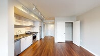 Located at 20 E Lanvale Street in Baltimore's midtown neighborhood, Unit 211 is a thoughtfully designed single-floor residence that blends modern finishes with functional living. Rich, dark hardwood flooring extends throughout the open-concept main living area, creating a warm yet contemporary aesthetic.

The kitchen is well-appointed with a full suite of stainless steel appliances, including an electric range with oven, refrigerator, and dishwasher. White flat-front cabinetry — including open upper shelving — pairs with dark countertops for a sleek, modern contrast. Track lighting overhead ensures the space is brightly and evenly lit. The kitchen flows seamlessly into the open living area, making the layout ideal for everyday living and entertaining.

The living area is generously proportioned and bathed in natural light from a large picture window offering views of the surrounding Baltimore cityscape. Neutral gray walls complement the warm flooring tones and provide a versatile backdrop.

A dedicated closet with a rod and shelving system offers practical storage, while a hallway leads to additional rooms. The bathroom features stone-look tile flooring, subway tile in the walk-in shower, a vanity with dark countertop, and brushed nickel hardware and towel bars, lending a refined, contemporary finish to the space.