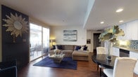 This well-appointed single-floor residence offers a thoughtfully designed open-concept layout that balances comfort and modern style. The living area serves as the heart of the home, centered around rich hardwood flooring and anchored by a large sectional sofa in warm neutral tones, complemented by deep navy blue accent pillows and a bold navy area rug. A sleek glass coffee table adds a contemporary touch, while a framed abstract artwork in soft blues and grays adorns the wall above, tying the color palette together cohesively. Recessed lighting and a smooth white ceiling with a subtle tray detail contribute to the clean, polished ambiance throughout. Large sliding glass doors flood the living space with abundant natural light and provide direct access to a private outdoor balcony, where outdoor seating is available for relaxing or dining al fresco. Adjacent to the living room, a round dining table with tufted white chairs offers an intimate dining setting. The kitchen is visible in the background, featuring white cabinetry and open shelving with wine storage, suggesting a practical yet refined culinary space. Overall, the residence presents a cohesive, modern aesthetic with warm tones and thoughtful finishes throughout.