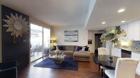 This well-appointed single-floor residence offers a thoughtfully designed open-concept layout that balances comfort and modern style. The living area serves as the heart of the home, centered around rich hardwood flooring and anchored by a large sectional sofa in warm neutral tones, complemented by deep navy blue accent pillows and a bold navy area rug. A sleek glass coffee table adds a contemporary touch, while a framed abstract artwork in soft blues and grays adorns the wall above, tying the color palette together cohesively. Recessed lighting and a smooth white ceiling with a subtle tray detail contribute to the clean, polished ambiance throughout. Large sliding glass doors flood the living space with abundant natural light and provide direct access to a private outdoor balcony, where outdoor seating is available for relaxing or dining al fresco. Adjacent to the living room, a round dining table with tufted white chairs offers an intimate dining setting. The kitchen is visible in the background, featuring white cabinetry and open shelving with wine storage, suggesting a practical yet refined culinary space. Overall, the residence presents a cohesive, modern aesthetic with warm tones and thoughtful finishes throughout.