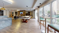 Located at 200 College, this single-floor amenity lounge is thoughtfully designed to provide a dynamic and social environment for its users. The space is anchored by a striking open layout with light wood flooring that flows seamlessly throughout, connecting each distinct zone with a cohesive aesthetic.

At the heart of the space sits a contemporary billiards table, positioned beneath pendant lighting and flanked by large floor-to-ceiling sliding glass doors that flood the area with natural light and offer views of the outdoor surroundings. A slatted wood partition wall serves as a stylish room divider, framing a wall-mounted flat-screen TV and lending an organic, architectural warmth to the interior.

Adjacent to the billiards area is a comfortable lounge zone featuring plush, modern armchairs arranged on a colorful geometric area rug, creating an inviting space for conversation or casual relaxation. A separate media lounge offers additional seating alongside a large mounted television, vibrant yellow accent walls, decorative artwork, and a warm, layered atmosphere.

The kitchenette is sleek and functional, outfitted with wood cabinetry, stainless steel appliances, white countertops, a sink, and dual wall-mounted TVs, alongside a chalkboard accent wall that adds a playful, communal touch. Recessed and track lighting throughout ensures each area is well-lit, while the overall design balances contemporary style with welcoming comfort.