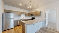 This thoughtfully designed two-bedroom, two-bathroom residence offers a modern and functional layout that makes excellent use of its single-floor footprint. The open-concept kitchen and living area serves as the heart of the home, anchored by a striking peninsula with granite countertops and geometric pendant lighting overhead. The kitchen is outfitted with rich taupe shaker-style cabinetry, a classic tile backsplash, and a full suite of stainless steel appliances, including a refrigerator, range, microwave, and dishwasher. Light wood-look flooring runs throughout the main living area, lending warmth and continuity to the space. Both bedrooms are carpeted for comfort, each featuring a ceiling fan, natural light from well-placed windows with blinds, and access to a dedicated closet with built-in shelving and hanging rods. The bathrooms are clean and modern, featuring wood-look tile flooring, granite-topped vanities with taupe cabinetry, and a full tub-shower combination. A carpeted staircase with a handrail leads to the entrance, adding a welcoming architectural touch. Neutral wall tones and recessed lighting throughout create a bright, cohesive ambiance well-suited for comfortable everyday living.