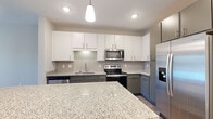 This well-appointed single-floor unit offers a contemporary design with a thoughtful layout that balances open living spaces with comfortable private areas. The kitchen serves as a central highlight, showcasing crisp white upper cabinetry paired with charcoal gray lower cabinets, a subway tile backsplash, and gleaming granite countertops. A generously sized center island provides additional prep space and casual seating, illuminated by elegant pendant lighting overhead. Stainless steel appliances, including a refrigerator with ice maker, electric range, microwave, and dishwasher, complete the modern culinary space. The kitchen flows seamlessly into the main living area, which features durable wood-look flooring that extends throughout the common spaces. A door with an adjacent window opens to an outdoor area, allowing natural light to fill the room and providing a connection to the exterior. The bedrooms are finished with plush carpet flooring and neutral-toned walls, offering a calm retreat. One bedroom includes a ceiling fan with light fixture and a window with views of the surrounding area. Additional rooms feature clean lines, ample wall space, and wood-look flooring. The overall aesthetic is modern and neutral, creating a versatile and move-in ready environment.
