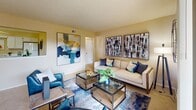 This single-floor residence offers a thoughtfully arranged layout with a warm, neutral color palette that creates a cohesive and inviting atmosphere throughout. The living room serves as the heart of the home, featuring soft wall-to-wall carpeting, a textured ceiling, and an open, generous footprint that comfortably accommodates a full seating arrangement. A distinctive pass-through architectural detail connects the living area to an adjacent space, adding visual interest and a sense of connectivity. Bold contemporary artwork accents the walls, complementing the modern furnishings that include a neutral sofa, navy accent chairs, and a glass-topped coffee table. The bedroom is generously sized, also finished with plush carpeting, and features a large upholstered headboard, ample natural light from a window with drape treatments, and mirrored wardrobe doors that enhance the sense of space. The kitchen is functional and bright, fitted with white cabinetry, white appliances including an electric range and over-the-range microwave, light countertops, and clean tile flooring. The bathroom offers a white vanity with a large mirror, tile flooring, a shower-tub combination, and warm overhead lighting, completing this well-maintained and move-in ready residence.