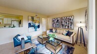 This single-floor residence offers a thoughtfully arranged layout with a warm, neutral color palette that creates a cohesive and inviting atmosphere throughout. The living room serves as the heart of the home, featuring soft wall-to-wall carpeting, a textured ceiling, and an open, generous footprint that comfortably accommodates a full seating arrangement. A distinctive pass-through architectural detail connects the living area to an adjacent space, adding visual interest and a sense of connectivity. Bold contemporary artwork accents the walls, complementing the modern furnishings that include a neutral sofa, navy accent chairs, and a glass-topped coffee table. The bedroom is generously sized, also finished with plush carpeting, and features a large upholstered headboard, ample natural light from a window with drape treatments, and mirrored wardrobe doors that enhance the sense of space. The kitchen is functional and bright, fitted with white cabinetry, white appliances including an electric range and over-the-range microwave, light countertops, and clean tile flooring. The bathroom offers a white vanity with a large mirror, tile flooring, a shower-tub combination, and warm overhead lighting, completing this well-maintained and move-in ready residence.