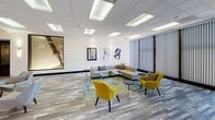 Located at 888 N Main St in Santa Ana, California, this sophisticated commercial lobby offers a refined and welcoming environment designed with both form and function in mind. The expansive open-plan floor is finished with wide-plank wood-look flooring in soft gray and beige tones, lending warmth and continuity throughout the space. A series of striking circular chandelier fixtures cascade along the ceiling, serving as a dramatic focal point that elevates the overall aesthetic. The main lounge area features a contemporary sectional sofa, vibrant yellow accent chairs, and a glass coffee table arranged atop a patterned area rug, creating an inviting gathering space bathed in light from large windows with vertical blinds. A dedicated seating nook near the entrance offers a pair of mustard leather chairs positioned to welcome visitors arriving through glass entry doors. A communal round table with modern chairs provides an additional meeting area, surrounded by curated abstract artwork on bright white walls and lush potted greenery. The elevator bank, clad in rich granite-toned stone with brass-accented doors, adds a classic, high-end touch. Recessed and panel ceiling lights ensure the space remains well-lit throughout, complementing the natural light that filters in from street-facing glazing. The overall ambiance is polished, professional, and contemporary.