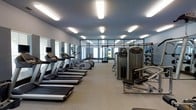 This modern fitness center is thoughtfully designed to accommodate a wide range of workout needs. The space is organized into distinct zones, with a row of treadmills and elliptical machines lining one side, providing ample cardio equipment for users. Opposite the cardio section, a selection of strength-training machines offers options for targeted muscle conditioning. Further into the space, a dedicated area houses free weights, medicine balls, and additional resistance equipment, catering to varied training styles.

The interior features a neutral color palette with light gray walls and flooring, contributing to a clean and focused atmosphere. Recessed rectangular lighting fixtures are evenly distributed across the ceiling, ensuring the space is brightly and uniformly lit. Full-length mirrors along one wall enhance the sense of openness while allowing users to monitor their form during exercise. Large glass partition walls at the rear of the room allow natural light to filter in and provide a visual connection to adjacent areas. The overall layout is spacious and well-organized, promoting an efficient and comfortable fitness experience.