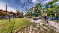 Located at 1250 Woodcrest Dr in Daytona Beach, Florida, this outdoor dog park offers a dedicated, fully enclosed space designed for pets and their owners to enjoy the Florida sunshine. The area is secured by a chain-link fence with a gated entry, ensuring a safe environment for dogs to roam and play freely.

The park features a mix of grassy turf and sandy ground, providing varied terrain that is natural and easy on paws. Agility equipment is a standout amenity, including a ramp and a tunnel-style obstacle, giving active dogs an engaging space to exercise and explore. Tall light poles are positioned throughout the enclosure, suggesting the space is usable during evening hours as well.

A shaded seating area with a picnic table and a large teal umbrella offers a comfortable spot for owners to relax while their pets play. The surrounding landscape is rich with native Florida vegetation, including towering palms, pine trees, and flowering shrubs, creating a lush, natural backdrop that enhances the overall ambiance of the space.

The park is situated adjacent to a residential building complex, making it a convenient on-site amenity. The open sky overhead, abundant natural light, and verdant surroundings contribute to a welcoming, resort-like atmosphere that is characteristic of the Daytona Beach region.