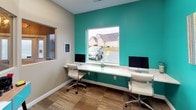 This single-floor commercial space makes a strong visual impression through its bold teal accent walls, which energize the interior and lend a modern, professional aesthetic throughout. The workstation area serves as the focal point of the space, featuring a continuous white countertop desk that accommodates multiple iMac computers side by side, paired with sleek white ergonomic chairs. A large picture window above the desk floods the workspace with natural light while offering views of the surrounding exterior. A pass-through window connects this area to an adjacent zone, enhancing the open, communicative layout of the space. The flooring transitions between warm wood-style planks and neutral carpet tile, adding texture and visual interest underfoot. A dedicated convenience corner features a teal-accented console table housing a printer and office supplies, with a prominent "WiFi" wall display signaling connectivity amenities for visitors. Artwork adorns the teal walls, including a large abstract canvas with vertical pastel and yellow tones, contributing to the creative and welcoming atmosphere. The entryway includes secured glass double doors with an illuminated exit sign, reinforcing the space's commercial functionality. Overall, the design balances practicality with personality, making it a functional and visually engaging environment suited for a business or service-oriented setting.
