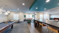 Located at 906 Cypress Rd in Saint Cloud, Minnesota, this single-floor community clubhouse is thoughtfully designed to offer a dynamic and welcoming social environment. The space features multiple distinct lounge zones, each furnished with colorful, contemporary seating including teal patterned armchairs, a bold striped curved sectional, and chevron-accented sofas — all accented by vibrant artwork displayed throughout. Recessed lighting illuminates the space consistently, complemented by natural light streaming through numerous windows.

A standout feature is the games area, anchored by a full-size pool table beneath a trio of elegant pendant lights, with an accent wall showcasing a geometric textured pattern. Nearby, casual high-top bar seating along a dark countertop provides an additional gathering spot, ideal for socializing or working. A tray ceiling detail with a bold teal painted inset adds architectural interest to the main lounge area.

The space also includes a kitchenette equipped with a refrigerator, dishwasher, and countertop coffee maker, making it convenient for events and gatherings. A recessed lounge nook with two sofas and bistro tables offers a more intimate setting. Overall, the clubhouse combines energetic color, functional amenities, and comfortable furnishings to create an engaging community hub.