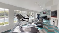 This well-appointed fitness center offers a bright and energizing environment designed to support a comprehensive workout experience. The space is flooded with natural light from a generous bank of large white-framed windows that line the walls, offering views of the surrounding outdoor amenities, including what appears to be a tennis court and lush greenery beyond.

The eye-catching chevron-patterned floor in teal, gray, and white adds a bold graphic element that energizes the space and gives it a modern, polished aesthetic. Crisp white walls and recessed lighting keep the atmosphere clean and open, while high ceilings contribute to an airy, spacious feel.

The fitness center is equipped with a well-curated selection of Matrix-brand cardio machines, including treadmills, ellipticals, and a rowing machine, arranged to maximize floor space and ease of movement. A dedicated strength training area features a multi-use cable machine and a rack of dumbbells, catering to a full range of resistance workouts. Mounted flat-screen televisions are positioned for easy viewing during cardio sessions.

A motivational typography accent wall in teal adds personality and visual interest to the strength training zone. Interior frosted-glass windows connect the fitness area to an adjacent space, allowing light to pass through while maintaining privacy. The overall design strikes a balance between function and style, making this fitness center both practical and visually compelling.