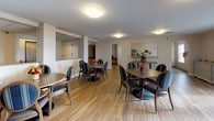 This communal gathering space is designed to accommodate multiple groups simultaneously, with several round and rectangular dining tables arranged across an open, airy floor plan. The warm hardwood-style flooring runs throughout, lending a welcoming and cohesive aesthetic to the entire area. Chairs upholstered in blue patterned and dark leather fabrics complement the wood-toned furniture, creating a comfortable yet orderly environment.

A notable architectural feature is the mezzanine-level balcony with white railings that overlooks the main floor, adding vertical interest and a sense of openness to the space. Flush-mount ceiling lights provide consistent, even illumination throughout. A wall-mounted flat-screen television is positioned for easy viewing from multiple seating areas, while a decorative console cabinet with ornate detailing adds a touch of elegance.

Pass-through service windows are integrated into interior walls, suggesting access to a service or kitchen area nearby. French doors along one wall allow natural light to enter and provide a connection to an exterior area beyond. Floral centerpieces on the tables contribute a cheerful, inviting atmosphere. A corridor visible from the main area leads to additional rooms, indicating this space serves as a central hub within a larger facility.