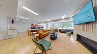 Located in Bloomfield, Connecticut, this thoughtfully designed single-floor community lounge space offers a dynamic and welcoming environment built around multiple distinct functional zones. Light hardwood flooring runs throughout, unifying the open layout and complementing the bright, well-lit interior illuminated by recessed linear ceiling fixtures and generous natural light from large windows overlooking lush greenery outside.

At the heart of the space, a central lounge area features a warm cognac leather sofa, a round coffee table, and an eclectic mix of green accent chairs arranged atop a woven geometric area rug. Adjacent to this is a standout fireplace wall clad in textured white geometric tile, topped by a large mounted flat-screen TV and flanked by vertical wood slat paneling — a striking focal point that anchors the room.

A semi-private nook showcases bold terracotta arch murals on the wall, housing high-backed tufted blue privacy sofas with chaise extensions, ideal for quiet conversation or focused work. Nearby, a communal dining and work table with contemporary chairs provides a collaborative space.

Toward the window-facing wall, counter-height bar seating and additional lounge sofas take full advantage of the outdoor views. A large photographic stone mural adds an artistic touch to one wall, while potted plants and carefully curated furnishings contribute to the space's lively yet comfortable ambiance.