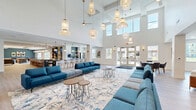 Located at 14744 Malachi Rd in Noblesville, Indiana, this impressive single-floor community clubhouse offers a generous and thoughtfully designed gathering space. The centerpiece is a breathtaking open-concept great room with soaring double-height ceilings, an abundance of natural light pouring in through clerestory windows, and a dramatic array of pendant and globe light fixtures suspended overhead. Expansive blue sectional sofas are arranged atop a large abstract area rug, creating an inviting and stylish social hub. French doors lead directly to an outdoor patio area, seamlessly blending indoor and outdoor living. The fully equipped kitchen features sleek dark granite countertops, white cabinetry, a built-in wine rack, stainless steel appliances, dual mounted televisions, and a long island with bar seating — ideal for hosting events. A cozy lounge area offers warm-toned walls, rich wood floating shelves, a linear fireplace feature wall, built-in cabinetry, and a wall-mounted display screen. A separate sitting room showcases linear recessed ceiling lighting, textured wall art, and plush upholstered seating. The welcoming entry lobby features light wood flooring, elevator access, mounted display screen, and bright glazed entry doors that flood the space with natural light.