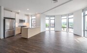 This contemporary 2-bedroom residence at 275 Washington Street in Newton, MA showcases a thoughtfully designed open-concept layout that seamlessly integrates the kitchen, dining, and living areas. Rich hardwood flooring extends throughout the entire unit, providing a warm contrast to the crisp white walls and ceilings.

The kitchen is a standout feature, anchored by a light wood island with a white quartz countertop and undermount sink. White cabinetry lines the walls, complemented by stainless steel appliances including a gas range, built-in microwave, and refrigerator. A window beside the range provides natural light and neighborhood views.

The living area is defined by a distinctive curved wall of floor-to-ceiling windows, flooding the space with abundant natural light and offering expansive views of the surrounding Newton streetscape. The generous open layout provides significant flexibility for furniture arrangement.

The bedrooms feature hardwood floors and large windows that continue the theme of natural light throughout. The bathroom is finished with a light wood vanity, white quartz countertop, a framed mirror, and a wall-mounted storage cabinet, reflecting the unit's clean, modern aesthetic.