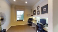 This single-floor business center is thoughtfully designed to offer a functional yet refined workspace. The room features two dedicated workstations, each equipped with a computer monitor, keyboard, and ergonomic office chair, arranged along a continuous built-in desk that lines the walls for an efficient use of space. Recessed lighting in the white ceiling provides consistent, even illumination throughout the room, complemented by a window fitted with horizontal blinds that invites natural light while maintaining privacy. The window ledge is accented with a small green plant arrangement, adding a touch of warmth to the professional setting. Crown molding frames the room with a classic architectural detail, while the neutral beige walls and carpeted flooring create a calm and focused atmosphere. The walls are adorned with a curated selection of framed artwork and decorative sunburst mirrors in metallic finishes, lending the space a stylish yet understated elegance. A tall decorative vase with dried branches adds a final organic touch. A posted notice near the workstation outlines business center guidelines, confirming the room's purpose as a shared professional resource.