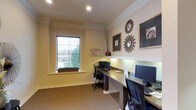 This single-floor business center is thoughtfully designed to offer a functional yet refined workspace. The room features two dedicated workstations, each equipped with a computer monitor, keyboard, and ergonomic office chair, arranged along a continuous built-in desk that lines the walls for an efficient use of space. Recessed lighting in the white ceiling provides consistent, even illumination throughout the room, complemented by a window fitted with horizontal blinds that invites natural light while maintaining privacy. The window ledge is accented with a small green plant arrangement, adding a touch of warmth to the professional setting. Crown molding frames the room with a classic architectural detail, while the neutral beige walls and carpeted flooring create a calm and focused atmosphere. The walls are adorned with a curated selection of framed artwork and decorative sunburst mirrors in metallic finishes, lending the space a stylish yet understated elegance. A tall decorative vase with dried branches adds a final organic touch. A posted notice near the workstation outlines business center guidelines, confirming the room's purpose as a shared professional resource.