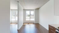 Located at 1115 NW Glisan St in Portland's vibrant Pearl District, this single-floor unit showcases a contemporary design aesthetic that blends industrial and modern elements throughout. Exposed concrete ceilings lend an urban character to the living areas, while rich hardwood flooring flows seamlessly from the open living space into the kitchen, creating a warm, cohesive interior.

The living area benefits from multiple windows that frame views of the surrounding Portland cityscape, allowing natural light to fill the space throughout the day. A partial partition wall adds a subtle architectural division without compromising the open, airy feel of the layout.

The kitchen is well-appointed with a clean, modern design featuring two-tone cabinetry — white upper cabinets paired with wood-grain lower cabinets — complemented by white quartz countertops and a glass tile backsplash. Stainless steel appliances, including a refrigerator, range, microwave, and dishwasher, complete the functional setup. A contemporary pendant chandelier adds a stylish focal point above the kitchen workspace.

The bathroom is finished with a wood-grain vanity, white countertop, and a full tub-shower combination, maintaining the clean, minimalist design language found throughout the rest of the unit.