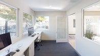 This single-floor business center is thoughtfully designed to provide a productive and comfortable workspace. The room features a clean, modern aesthetic with crisp white walls accented by a bold yellow feature wall adorned with a colorful piece of abstract artwork, adding a vibrant and creative energy to the space. Dark-toned carpet tiles ground the interior, offering a professional contrast to the bright surroundings. A long, built-in white workstation runs along the perimeter of the room, accommodating multiple desktop computer setups equipped with monitors, keyboards, and mice. Ample under-counter cabinetry provides convenient storage. A large multifunction printer and copier is stationed within easy reach, supporting a range of document needs. Large windows line the walls, inviting generous natural light and offering views of the surrounding outdoor greenery and neighborhood. A frosted glass door with etched signage serves as the entrance, maintaining privacy while preserving an open, airy feel. Recessed ceiling lighting complements the natural light, ensuring a well-lit environment throughout the day. Overall, this business center delivers a functional, well-appointed setting ideal for printing, computing, and focused work tasks.