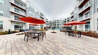This outdoor amenity space is set within a sweeping, multi-story residential complex and designed to serve as a central hub for relaxation and socializing. The centerpiece is a generously sized swimming pool with crystal-clear blue water, framed by a concrete pool deck lined with lounge chairs and bold red market umbrellas that provide ample shade throughout the day. An accessibility lift is present at the pool's edge, ensuring the space is welcoming to all residents.

Beyond the pool, an expansive courtyard features a broad paver patio laid in a classic pattern, furnished with dining-height tables and chairs, each shaded by signature red umbrellas. Curved, colorful outdoor sofas are thoughtfully positioned to create intimate conversation areas, while a separate garden nook offers lattice-style lounge chairs surrounded by seasonal plantings and manicured landscape beds.

Winding paver pathways connect the various seating zones, guiding visitors through the lush greenery and young trees that punctuate the courtyard with natural texture. The surrounding multi-story building facades, clad in neutral siding with red accent trim and featuring private balconies, enclose the space to create a sense of privacy and community. The overall ambiance is modern, inviting, and thoughtfully designed for outdoor living.