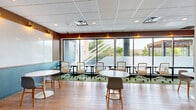 Located at 2508 SE Delaware St in Minneapolis, MN, this single-floor commercial space offers a thoughtfully designed collaborative environment. The layout comprises two distinct areas: an open coworking lounge and a semi-private office, connected by a stylish sliding barn door with frosted glass panels that allows for flexible use and easy transitions between spaces.

The main coworking area is bright and welcoming, lined with expansive floor-to-ceiling windows that flood the space with natural light and offer views of the surrounding greenery. The interior features warm wood-toned flooring throughout, accented by a rich teal accent wall and warm terracotta-brown upper trim. Seating is varied and functional, with round white pedestal tables paired with modern chairs for casual collaboration, as well as long workbench-style desks along the window wall outfitted with ergonomic mesh office chairs.

A standout feature is a dedicated whiteboard wall framed by bold yellow accents and illuminated by three distinctive cage-style pendant lights, creating an ideal zone for brainstorming and presentations. Vertical wood slat paneling adds architectural texture, while gold wall sconces provide warm ambient lighting throughout.

The private office, accessible through the sliding barn door, includes a dedicated desk and artwork, offering a quieter setting for focused work. The overall ambiance is energetic yet professional, combining colorful design elements with practical, flexible furnishings suited for a dynamic work environment.