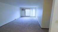 This single-floor space features an open living area finished with wall-to-wall carpeting in a neutral tone, contributing to a clean and functional layout. The room is brightened by a large sliding glass door fitted with vertical blinds, which floods the interior with natural light and provides direct access to an outdoor balcony. A wall-mounted air conditioning unit sits adjacent to the sliding door, offering climate control convenience. Baseboard heating runs along the perimeter of the room, ensuring year-round comfort. The walls are painted in a crisp white, enhancing the sense of openness throughout the space. The layout is straightforward and versatile, allowing for flexible furniture arrangement. Overall, the space presents a simple, practical design with key features that support comfortable everyday living.