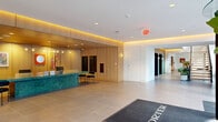 Located at 1105 Pleasant St in Oak Park, Illinois, The Porter presents a thoughtfully designed ground-floor lobby and common area that makes a strong first impression. Visitors are welcomed through a sleek glass revolving entrance door, stepping onto large-format porcelain tile flooring beneath high ceilings with recessed lighting. A standout feature of the lobby is the bold, curved reception desk crafted from richly veined green marble, set against a warm natural wood-paneled backdrop with integrated cove lighting that casts a welcoming amber glow. Curated artwork adorns the walls, adding a vibrant, contemporary character to the space. Adjacent to the reception area, a comfortable lounge seating area with modern furnishings provides a relaxed gathering spot for residents and guests. A dramatic open-riser staircase with black metal railings and wood treads rises along a dramatic floor-to-ceiling window wall, allowing generous natural light to pour in and offering views of the lively Oak Park streetscape. Lush potted plants positioned throughout the corridor enhance the biophilic design aesthetic. The space also includes a well-organized back-of-house utility room with ample built-in shelving, supporting the functional needs of the building's operations. Overall, the design balances refined materials with a warm, modern sensibility.
