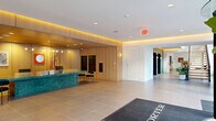 Located at 1105 Pleasant St in Oak Park, Illinois, The Porter presents a thoughtfully designed ground-floor lobby and common area that makes a strong first impression. Visitors are welcomed through a sleek glass revolving entrance door, stepping onto large-format porcelain tile flooring beneath high ceilings with recessed lighting. A standout feature of the lobby is the bold, curved reception desk crafted from richly veined green marble, set against a warm natural wood-paneled backdrop with integrated cove lighting that casts a welcoming amber glow. Curated artwork adorns the walls, adding a vibrant, contemporary character to the space. Adjacent to the reception area, a comfortable lounge seating area with modern furnishings provides a relaxed gathering spot for residents and guests. A dramatic open-riser staircase with black metal railings and wood treads rises along a dramatic floor-to-ceiling window wall, allowing generous natural light to pour in and offering views of the lively Oak Park streetscape. Lush potted plants positioned throughout the corridor enhance the biophilic design aesthetic. The space also includes a well-organized back-of-house utility room with ample built-in shelving, supporting the functional needs of the building's operations. Overall, the design balances refined materials with a warm, modern sensibility.