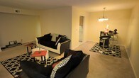 This single-floor residence offers a comfortable and cohesive layout designed for everyday living. The living area is generously sized, centered around bold black upholstered sofas accented with black-and-white patterned throw pillows, creating a striking visual contrast against the warm, neutral-toned walls and plush carpeted flooring. A geometric-patterned area rug anchors the seating arrangement, while a compact side table adds a functional touch to the space.

The living area flows seamlessly into the dining space, which is distinguished by an elegant chandelier overhead, casting a warm glow across the room. A raised bar-height table with matching stools provides a stylish dining setup, complemented by a dark accent cabinet nearby.

Adjacent to the main living space, the kitchen is visible through an open passageway, featuring cabinetry and standard appliances that make it both practical and accessible. The consistent use of soft, warm paint tones throughout the space creates a unified and inviting ambiance.

Overall, this residence balances functional design with tasteful décor, offering a welcoming environment that is both practical and visually cohesive across its open, connected living areas.