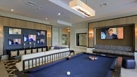 This single-floor entertainment lounge is thoughtfully designed to provide a premium social and recreational experience. At its centerpiece sits a full-size pool table with a vibrant blue felt surface, inviting guests to enjoy a game in a sophisticated setting. Flanking the pool table are sleek, modern sofas and seating arrangements upholstered in neutral tones, offering comfortable spots for lounging and conversation.

The walls are adorned with large, framed flat-screen televisions, making this an ideal space for watching live events or catching up on the latest news. Decorative wall sconces and a striking rectangular pendant light fixture overhead—accented with natural branch-like elements—add warmth and visual interest to the room.

The interior design balances contemporary and refined aesthetics, featuring rich wood-paneled wall accents, large mirror panels that enhance the sense of space, and a carefully curated color palette of neutral creams, blacks, and warm tones. Recessed lighting complements the ambient fixtures, ensuring the room is well-lit throughout. An elevator visible in the background hints at this space being part of a larger multi-use facility. Overall, the lounge exudes an upscale, welcoming atmosphere suited for both casual recreation and social gatherings.