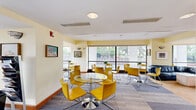 This single-floor communal space combines a kitchen and living area into one well-appointed, open-plan layout. The interior is defined by an expansive wall of floor-to-ceiling windows that runs along the perimeter, flooding the space with natural light and offering unobstructed views of surrounding greenery. Warm yellow walls complement the vibrant yellow upholstered chairs and glass-topped round tables, creating a lively and welcoming atmosphere throughout the lounge area. Multiple seating arrangements are distributed across the space, accommodating both small gatherings and larger groups with ease. A navy blue sectional sofa anchors one corner of the room, providing a comfortable lounge area alongside a flat-screen television mounted on a warm wood-paneled wall. A textured area rug adds visual depth beneath the seating zone. The kitchen features a curved granite-topped counter finished in deep blue cabinetry, with bar-height yellow stools lined along its length. Track lighting highlights the counter area, while framed artwork decorates the walls throughout, adding a curated, personalized touch. The flooring consists of a patterned carpet tile that ties the varied zones together cohesively. Overall, the space exudes a bright, modern, and social ambiance suited to community gathering and everyday use.