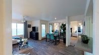 This single-floor residence offers an open-concept layout that connects the living, dining, and sleeping areas in a cohesive and functional design. The living area features rich, dark wood-look flooring that extends throughout the main space, creating a warm and grounded aesthetic. A ceiling fan with a classic white finish adds both style and practicality overhead. Natural light filters in through a glass-paneled door, brightening the room and providing a view to the outdoor area beyond. The dining space is defined by a sleek dark table surrounded by vibrant teal upholstered chairs, adding a bold pop of color to the otherwise neutral palette. A striking set of arched mirrored panels serves as an elegant room divider, reflecting light and adding depth to the space while maintaining an open feel. A large fiddle-leaf fig plant anchors one corner, bringing a touch of nature indoors. The adjacent bedroom, visible through the open layout, features plush carpeting and bright white furnishings, offering a calm and restful retreat. The overall ambiance is contemporary and inviting, with a carefully curated mix of textures, colors, and materials that create a polished, move-in-ready feel throughout.
