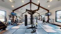This fitness center is housed within a striking single-story structure distinguished by its soaring vaulted ceilings accented with bold, dark-stained exposed wood beams and trusses. Recessed lighting and pendant fixtures illuminate the expansive interior, complementing the generous natural light that pours in through large black-framed windows positioned along multiple walls. Views of an outdoor pool area are visible from select windows, enhancing the open, airy atmosphere.

The flooring features a geometric patchwork design in varying shades of blue and neutral tones, adding a modern, polished aesthetic to the space. Equipment is thoughtfully distributed throughout, including a row of treadmalls and ellipticals for cardiovascular training, a multi-station cable machine, a Magnolia cable system, and an Instinct functional trainer. A central heavy bag and TRX suspension station serve as a focal point for functional and combat-style training. A dedicated free weight area includes a dumbbell rack and weight bench.

A decorative geometric lattice panel serves as an architectural accent, dividing zones within the facility. Multiple wall-mounted televisions are positioned throughout for entertainment. The overall ambiance is modern yet warm, blending rustic architectural details with contemporary fitness amenities.