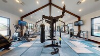 This fitness center is housed within a striking single-story structure distinguished by its soaring vaulted ceilings accented with bold, dark-stained exposed wood beams and trusses. Recessed lighting and pendant fixtures illuminate the expansive interior, complementing the generous natural light that pours in through large black-framed windows positioned along multiple walls. Views of an outdoor pool area are visible from select windows, enhancing the open, airy atmosphere.

The flooring features a geometric patchwork design in varying shades of blue and neutral tones, adding a modern, polished aesthetic to the space. Equipment is thoughtfully distributed throughout, including a row of treadmalls and ellipticals for cardiovascular training, a multi-station cable machine, a Magnolia cable system, and an Instinct functional trainer. A central heavy bag and TRX suspension station serve as a focal point for functional and combat-style training. A dedicated free weight area includes a dumbbell rack and weight bench.

A decorative geometric lattice panel serves as an architectural accent, dividing zones within the facility. Multiple wall-mounted televisions are positioned throughout for entertainment. The overall ambiance is modern yet warm, blending rustic architectural details with contemporary fitness amenities.