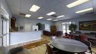 This single-floor space is designed to serve multiple functions within an open, well-lit layout. The interior features a drop ceiling equipped with recessed fluorescent panel lighting that evenly illuminates the entire area, complementing the light hardwood flooring that runs throughout the space.

A reception or service counter near the entrance provides a practical focal point, finished with a granite-style surface and backed by a whiteboard and an informational display board. Adjacent to the counter, a wooden armoire adds a traditional accent to the otherwise contemporary setting.

The lounge area is furnished with deep burgundy upholstered armchairs and a low coffee table, creating a comfortable seating arrangement ideal for informal gatherings or relaxed conversation. Nearby, large windows allow natural light to filter in, offering views of the outdoors and enhancing the overall brightness of the space.

A dining or meeting area occupies another section of the room, centered around a large round table surrounded by matching burgundy chairs, accommodating group activities or meals. Potted plants are strategically placed throughout, adding warmth and a natural element to the functional, community-oriented environment.