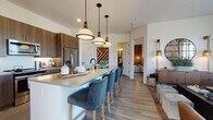 This thoughtfully designed 2-bedroom, 2-bathroom single-floor residence blends contemporary style with warm, inviting finishes throughout. The open-concept kitchen is a standout feature, showcasing rich dark wood cabinetry, stainless steel appliances, and a generous quartz-topped island with seating for multiple guests, illuminated by a trio of striking pendant lights with brass accents. Light wood-style flooring flows seamlessly through the main living areas, tying the spaces together cohesively.

The dining area is bathed in natural light from floor-to-ceiling windows and a glass door that opens to the outdoors, offering open exterior views. The living room continues this bright, airy feel with large windows framing outdoor scenery and providing a comfortable, welcoming atmosphere.

The primary bedroom features plush carpeting, generous natural light, and direct access to an en-suite bathroom appointed with granite countertops, dark wood vanity cabinetry, a glass-enclosed walk-in shower, and stylish tile flooring. The second bedroom is equally well-lit, with soft carpeting and ample natural light. A second full bathroom includes a soaking tub with subway tile surround and convenient access to a walk-in closet, delivering practical storage solutions throughout this refined residence.