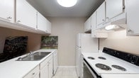 This single-floor residence offers a thoughtfully laid-out living environment designed for comfort and practicality. The kitchen is a standout feature, designed in a classic galley configuration that maximizes efficiency within its compact footprint. White shaker-style cabinetry lines both walls from floor to ceiling, providing ample storage space. The countertops are finished in a clean white surface, complementing the bright, cohesive aesthetic. A stainless steel double-basin sink is positioned beneath the upper cabinets on one side, while the opposite wall accommodates a full-size white refrigerator, a built-in dishwasher, and an electric coil range with overhead ventilation. The light tile flooring adds a subtle texture underfoot and reflects the overhead flush-mount ceiling light, keeping the space feeling bright and open. Neutral taupe-toned walls run throughout, creating a calm and versatile backdrop. A decorative painting adds a touch of warmth and character to the kitchen's end wall. The textured ceiling is a notable architectural detail consistent across the unit. Overall, the space presents a clean, functional, and welcoming environment well-suited for everyday living.