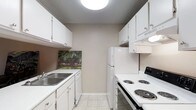 This single-floor residence offers a thoughtfully laid-out living environment designed for comfort and practicality. The kitchen is a standout feature, designed in a classic galley configuration that maximizes efficiency within its compact footprint. White shaker-style cabinetry lines both walls from floor to ceiling, providing ample storage space. The countertops are finished in a clean white surface, complementing the bright, cohesive aesthetic. A stainless steel double-basin sink is positioned beneath the upper cabinets on one side, while the opposite wall accommodates a full-size white refrigerator, a built-in dishwasher, and an electric coil range with overhead ventilation. The light tile flooring adds a subtle texture underfoot and reflects the overhead flush-mount ceiling light, keeping the space feeling bright and open. Neutral taupe-toned walls run throughout, creating a calm and versatile backdrop. A decorative painting adds a touch of warmth and character to the kitchen's end wall. The textured ceiling is a notable architectural detail consistent across the unit. Overall, the space presents a clean, functional, and welcoming environment well-suited for everyday living.