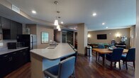 The Club Room is a well-appointed community gathering space designed to accommodate a variety of social activities. Rich, dark hardwood flooring flows throughout the entire space, anchoring the warm, neutral taupe and gray color palette. The kitchen area serves as a central hub, featuring dark cabinetry with ample storage, light quartz countertops, stainless steel appliances including a full-sized refrigerator and microwave, and a spacious island with seating for multiple bar-height chairs. Pendant lighting hangs above the island, adding focused illumination and a modern aesthetic touch. Recessed ceiling lights provide consistent brightness throughout.

Adjacent to the kitchen, a game and entertainment zone includes a full-sized billiards table and a wall-mounted flat-screen television, making it ideal for casual recreation. A dedicated lounge area offers generous natural light through large windows with views of the surrounding grounds. This seating zone features a plush sofa, coordinating blue accent chairs, a wood coffee table, and a patterned area rug, creating a comfortable and inviting retreat. A secondary seating arrangement with a wall-mounted TV and a dining table with blue chairs provides additional gathering options. The overall layout supports both lively group events and relaxed everyday use.