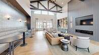 Located at 1120 Corner Wind Ln in Columbia, SC, this single-floor community space is a standout example of modern yet warm architectural design. The interior is anchored by dramatic vaulted ceilings adorned with striking exposed dark wood truss beams, creating an impressive sense of verticality and grandeur throughout. Large arched and rectangular windows line the walls, flooding the space with abundant natural light and offering views of the surrounding outdoor landscape.

Warm-toned hardwood flooring runs throughout, tying together the open, flowing layout seamlessly. The main living area features a generously sized lounge zone with plush seating arrangements, including a tan leather sofa, accent chairs, and a central coffee table, all set atop a neutral area rug. A built-in shelving unit with sage green cabinetry and a wet bar serves as a functional and stylish focal point, complemented by decorative objects and greenery.

A dedicated café-style nook offers bistro seating alongside a coffee station, ideal for casual gatherings. Toward the entry, a separate seating cluster with leather armchairs is framed by tall potted plants and sleek barn-style sliding doors. A staircase with a bold geometric accent wall adds a distinctive architectural touch. The overall ambiance is polished, welcoming, and thoughtfully curated for community use.
