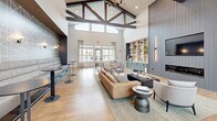 Located at 1120 Corner Wind Ln in Columbia, SC, this single-floor community space is a standout example of modern yet warm architectural design. The interior is anchored by dramatic vaulted ceilings adorned with striking exposed dark wood truss beams, creating an impressive sense of verticality and grandeur throughout. Large arched and rectangular windows line the walls, flooding the space with abundant natural light and offering views of the surrounding outdoor landscape.

Warm-toned hardwood flooring runs throughout, tying together the open, flowing layout seamlessly. The main living area features a generously sized lounge zone with plush seating arrangements, including a tan leather sofa, accent chairs, and a central coffee table, all set atop a neutral area rug. A built-in shelving unit with sage green cabinetry and a wet bar serves as a functional and stylish focal point, complemented by decorative objects and greenery.

A dedicated café-style nook offers bistro seating alongside a coffee station, ideal for casual gatherings. Toward the entry, a separate seating cluster with leather armchairs is framed by tall potted plants and sleek barn-style sliding doors. A staircase with a bold geometric accent wall adds a distinctive architectural touch. The overall ambiance is polished, welcoming, and thoughtfully curated for community use.