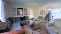 This single-floor space offers a warm and inviting atmosphere, anchored by a comfortably furnished living area designed for relaxed everyday living. The living room showcases a large sectional sofa adorned with decorative pillows, paired with a plush armchair, creating an ideal arrangement for conversation and lounging. A dark-finished coffee table serves as the centerpiece of the seating area, accessorized with a sculptural décor piece that adds a touch of personality. Soft carpeting underfoot enhances the room's cozy ambiance, while warm lamp lighting contributes to an intimate, welcoming feel. The walls are tastefully decorated with a framed mirror flanked by circular wall art panels, alongside a vibrant floral canvas print, giving the space a curated yet comfortable character. A dining area is visible in the background, featuring a bistro-style table and chairs, seamlessly extending the living space's functionality. The open layout connects these areas naturally, promoting an easy flow throughout the floor. Overall, the space balances practicality and style, offering a comfortable and well-organized environment suitable for day-to-day living.