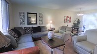 This single-floor space offers a warm and inviting atmosphere, anchored by a comfortably furnished living area designed for relaxed everyday living. The living room showcases a large sectional sofa adorned with decorative pillows, paired with a plush armchair, creating an ideal arrangement for conversation and lounging. A dark-finished coffee table serves as the centerpiece of the seating area, accessorized with a sculptural décor piece that adds a touch of personality. Soft carpeting underfoot enhances the room's cozy ambiance, while warm lamp lighting contributes to an intimate, welcoming feel. The walls are tastefully decorated with a framed mirror flanked by circular wall art panels, alongside a vibrant floral canvas print, giving the space a curated yet comfortable character. A dining area is visible in the background, featuring a bistro-style table and chairs, seamlessly extending the living space's functionality. The open layout connects these areas naturally, promoting an easy flow throughout the floor. Overall, the space balances practicality and style, offering a comfortable and well-organized environment suitable for day-to-day living.