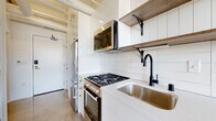 This single-floor unit blends industrial character with contemporary finishes throughout its well-defined layout. The kitchen is thoughtfully equipped with stainless steel appliances, including a gas range with oven, an over-the-range microwave, and a full-size refrigerator. White cabinetry pairs with open wood shelving mounted on black metal brackets, while a white tile backsplash and light quartz countertops create a clean, modern aesthetic. A matte black pull-down faucet and undermount stainless steel sink serve as refined focal points against the bright cabinetry.

The bathroom features polished concrete flooring, a wood-toned vanity with an undermount sink, and clean white walls that create a minimalist, functional space. A wall-mounted medicine cabinet provides additional storage.

The living and sleeping areas showcase polished concrete floors that reflect natural light, contributing to the unit's bright and open feel. Exposed painted brick along the lower walls adds texture and industrial character, while large, multi-pane black-framed windows frame urban skyline views and allow abundant natural light. A wall-mounted mini-split unit provides climate control. The overall ambiance is modern and urban, with a balance of raw industrial elements and refined contemporary finishes.