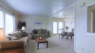 This single-story residence features an open-plan layout that seamlessly connects the living and dining areas, creating a spacious and cohesive environment. The living area is anchored by plush, neutral-toned sofas accented with patterned throw pillows, complemented by a classic wooden coffee table that adds warmth to the space. A framed artwork piece adorns the wall, enhancing the room's understated aesthetic. The textured ceiling, trimmed with crown molding, adds architectural character overhead, while a ceiling fan serves the dining area, providing both function and style. The dining space accommodates a glass-top table surrounded by sleek black chairs, with a floral centerpiece adding a touch of color. French doors along the back wall flood the space with natural light and offer a connection to the outdoors. Soft, wall-to-wall carpeting in a warm beige tone runs throughout, tying the two areas together cohesively. The overall color palette is light and neutral, with cream-colored walls contributing to a bright and welcoming ambiance suited for comfortable everyday living.