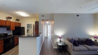 This single-floor Dallas residence offers a well-organized, open-concept layout designed for comfortable everyday living. Dark hardwood flooring runs throughout the main living areas, providing a warm and cohesive aesthetic that ties the space together.

The kitchen is positioned at the front of the main living zone, featuring a raised breakfast bar with dark granite-style countertops — an ideal spot for casual dining or meal prep. Rich, dark wood cabinetry adds depth and contrast, while a stainless steel refrigerator and sink complete the functional workspace.

Adjacent to the kitchen, the living area offers an inviting atmosphere with neutral gray walls and ample room for seating. A small accent table with a lamp adds a cozy touch to the corner, enhancing the warm ambiance of the space.

A central hallway connects the living areas to the private spaces deeper within the home, with natural light visible at the far end, hinting at well-lit rooms beyond. The corridor is clean and open, contributing to the overall sense of flow and accessibility throughout the layout.

The neutral color palette of whites and soft grays, combined with flush-mount ceiling lighting, creates a bright, understated interior that is both practical and inviting — well-suited for a range of furnishing styles.