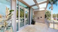 This single-floor space presents two distinct yet equally inviting entry experiences that set the tone for the interior. The first entrance features a sleek, contemporary design with floor-to-ceiling aluminum-framed glass doors and panels, allowing an abundance of natural light to flood the entryway. A translucent overhead canopy further enhances the bright, open atmosphere, while natural stone tile flooring and a textured stone accent wall add warmth and grounding to the modern aesthetic. Potted tropical plants provide a welcoming touch of greenery.

The second entrance takes on a more classic architectural character, highlighted by a teal fabric awning, warm wood-toned glass-paneled doors, and a decorative white balustrade railing. Tall, dark ceramic planters with lush ornamental grasses flank the entryway, adding elegance and curb appeal. The surrounding multi-story building exterior features terracotta tones, wrought iron balconies, and mature palm and cypress trees, creating a Mediterranean-inspired ambiance. Together, both entrances reflect a space that balances modern functionality with timeless design, offering a polished and professional first impression.