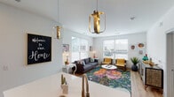This single-floor residence offers a thoughtfully designed open-concept layout that seamlessly connects the kitchen, dining, and living areas. The kitchen is outfitted with stainless steel appliances, grey cabinetry, a white quartz island with bar seating, and light hardwood flooring that extends throughout the main living spaces. Pendant lighting adds a warm, stylish accent above both the island and the living area. The living room is bright and inviting, benefiting from multiple large windows that fill the space with natural light. An exterior door off the kitchen provides convenient outdoor access. The primary bedroom features plush carpeting and a rustic wood headboard complemented by warm-toned nightstands, creating a cozy, mid-century modern atmosphere. The second bedroom is well-proportioned, also carpeted, and includes a window with natural light and ample wall space. The bathroom is clean and functional, featuring light wood-look tile flooring, a single vanity, and a tub-shower combination. Throughout the residence, crisp white walls and a cohesive design palette create a fresh, modern ambiance, while eclectic and colorful décor accents lend the space a lively, welcoming personality.